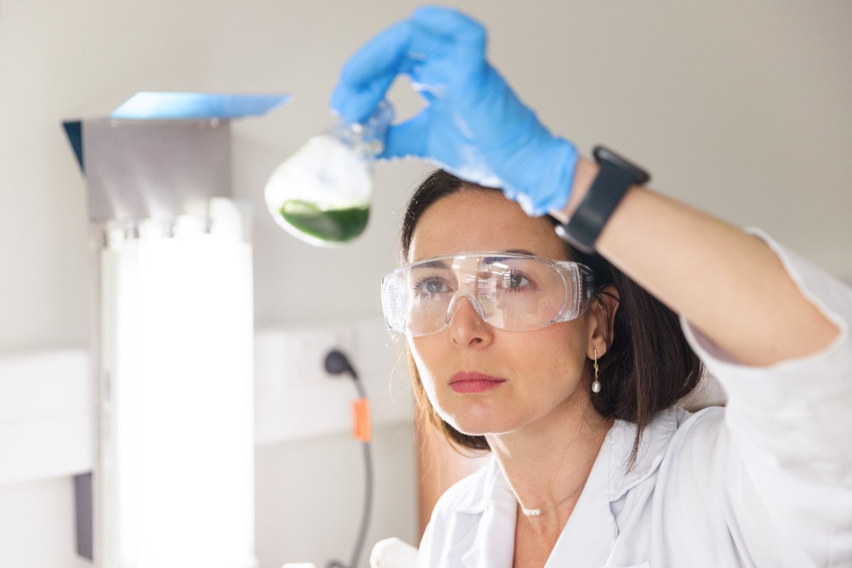 Photo of Dr Caroline Thum working in a laboratory. The image is framed showing their head and shoulders. They are wearing safety glasses, gloves, and a lab-coat, and holding up a glass beaker containing a green liquid.